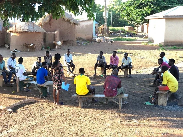 People sitting in a circle under a tree listening to a speaker